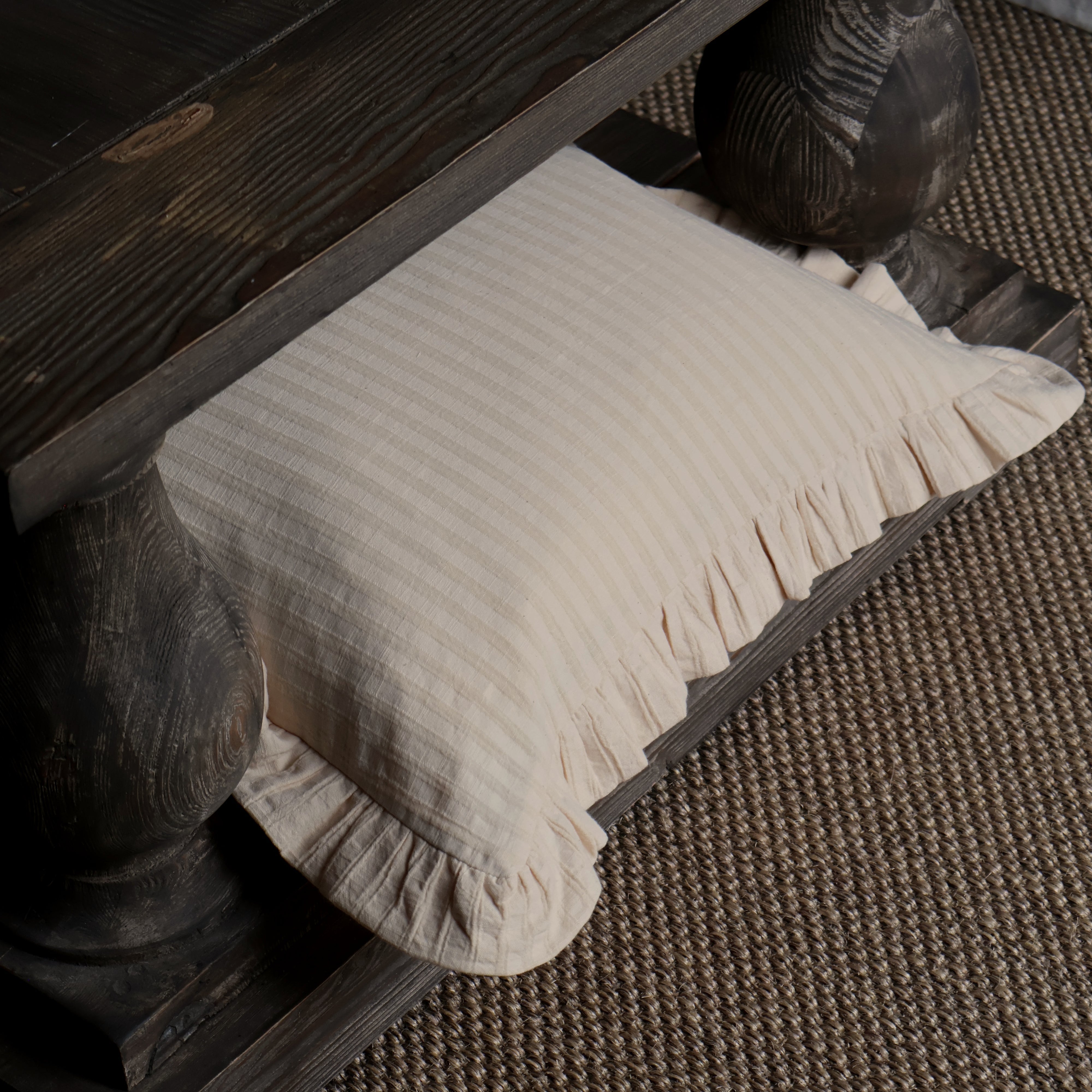 A cream-colored linen throw pillow with a ruffled edge, styled under a dark rustic wooden table on a textured brown and beige woven rug.