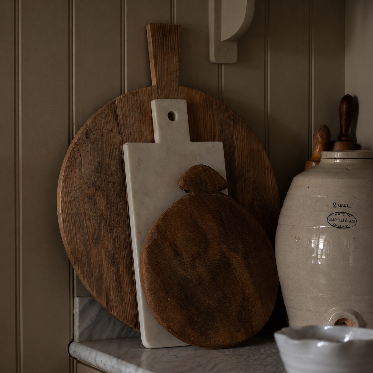 A rustic kitchen scene featuring wooden and stone cutting boards leaning against a paneled wall next to a large cream-colored ceramic crock pot.